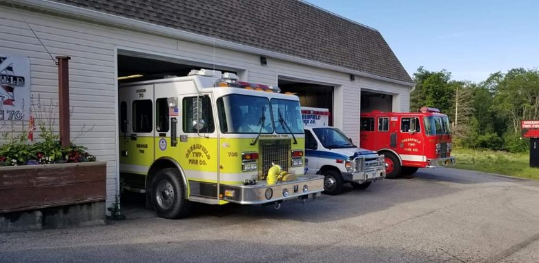 A white fire house showing open bay doors and three apparatus, one yellow fire rescue truck, one white and blue ambulance and a red fire engine.