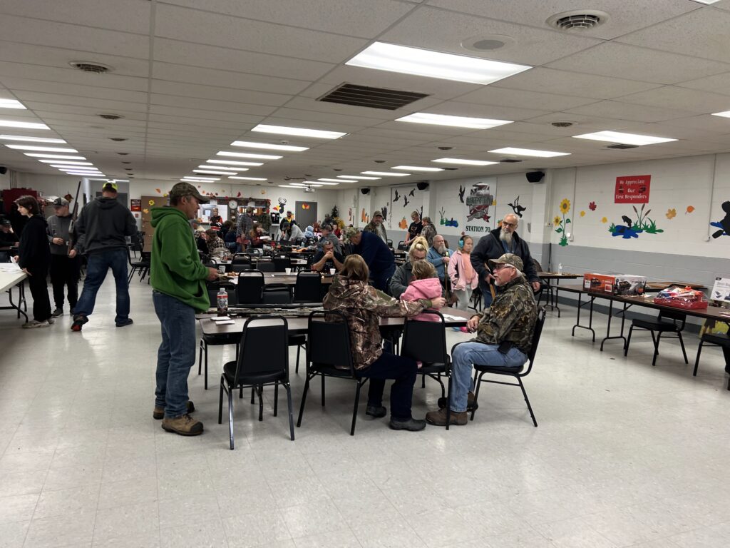 people sitting at tables is a large room, some standing and chatting, some sitting and chatting.