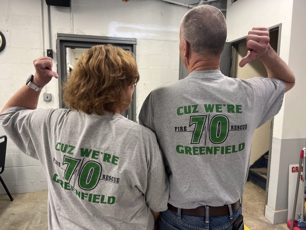 two individuals standing with their backs to the camera, one appears shorter and female, one taller and male with their arms raised and thumbs pointing to the backs of their shirts that read "cuz we're greenfield, 70"