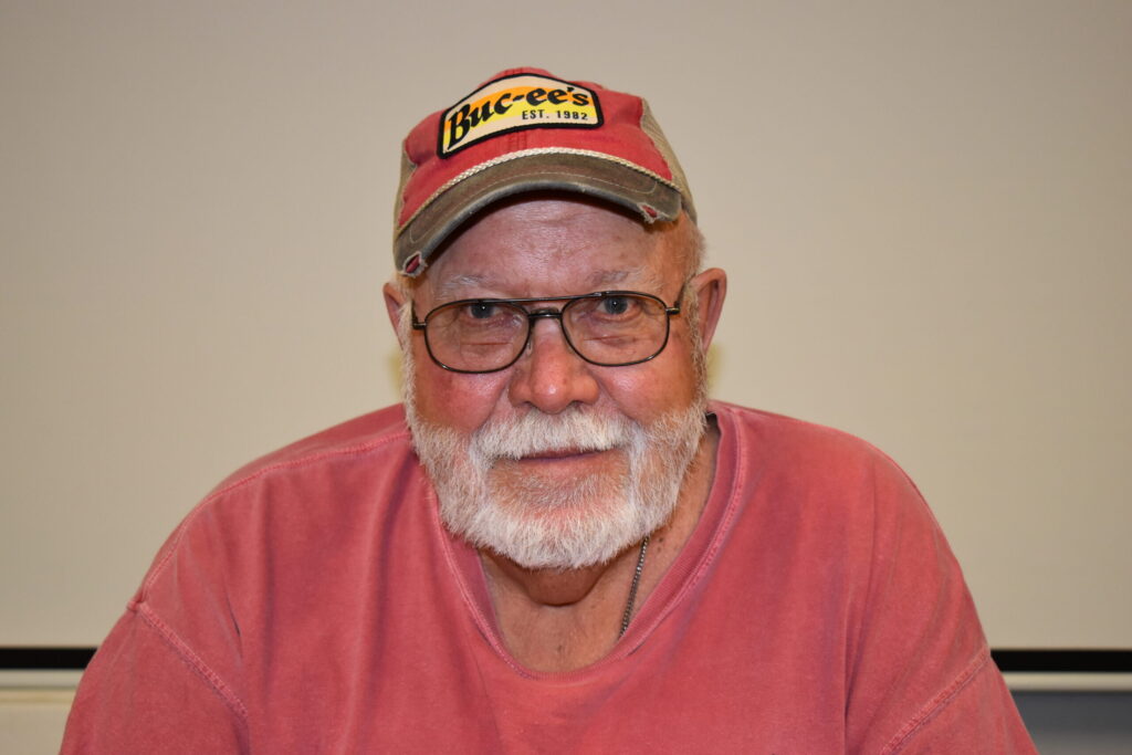 A man with a red shirt, a red and tan baseball cap, glasses, a beard and a mustache