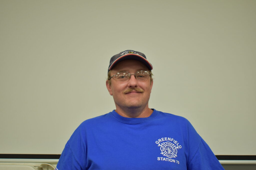 Man in bright blue shirt, dark blue hat and glasses with a mustache